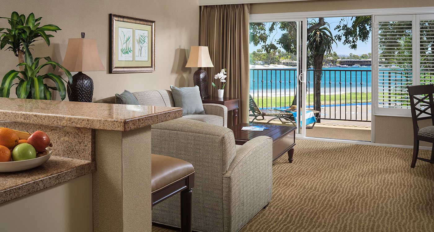 A wide view of a hotel suite with a living area, kitchenette, and balcony overlooking the ocean.