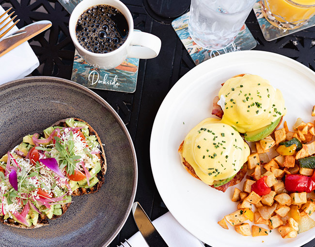 Overhead view of an elevated weekend brunch at Dockside 1953, showcasing fresh avocado toast topped with microgreens alongside eggs benedict, home fries, and hot morning coffee on the waterfront patio.