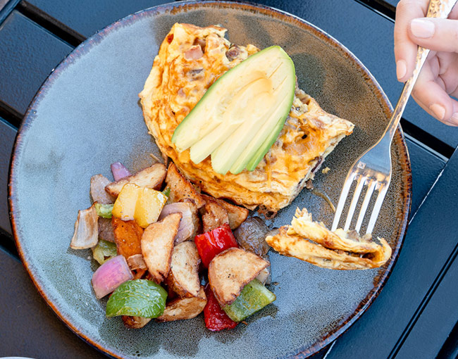 A freshly prepared breakfast omelet topped with sliced avocado, served with seasoned roasted potatoes and bell peppers on a waterfront patio dining table during weekend brunch at Dockside 1953.