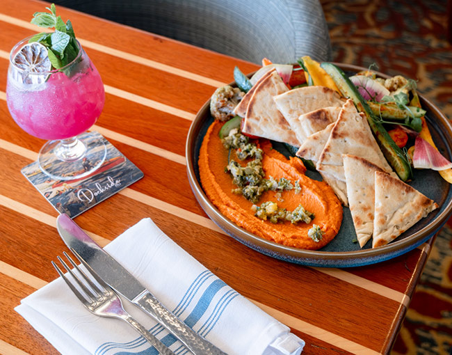 A premium California-coastal Hummus Plate served as an artisan shareable during weekend brunch on the waterfront patio at Dockside 1953, featuring smooth Edamame Hummus topped with roasted red pepper relish, crudité and warm grilled naan bread.