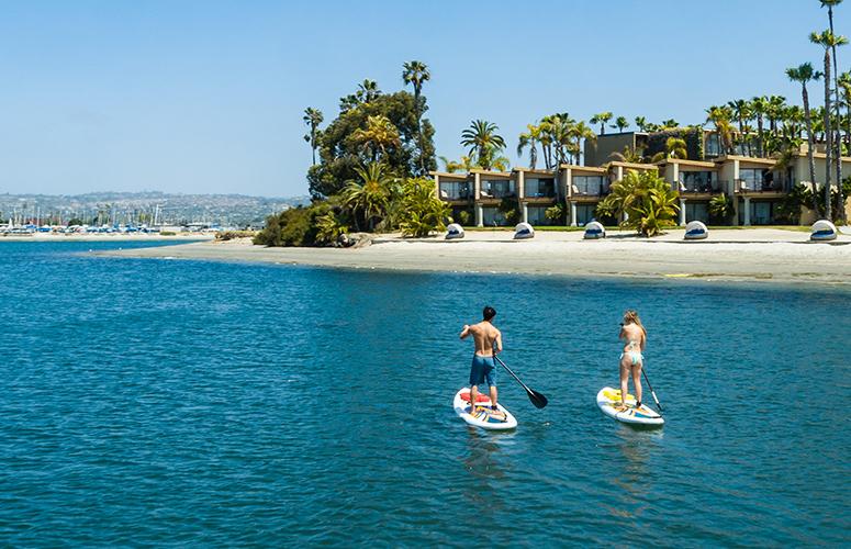 A beach scene with two people on paddleboards in the water and a hotel or resort in the background.